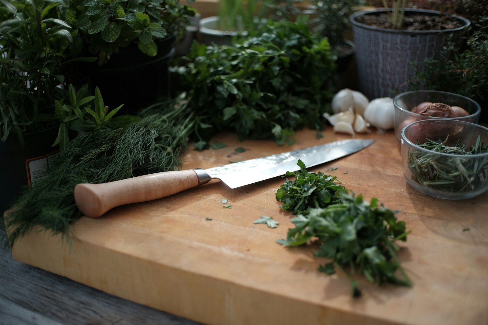 A chef's knife on a wooden board beside fresh chopped herbs and garlic
